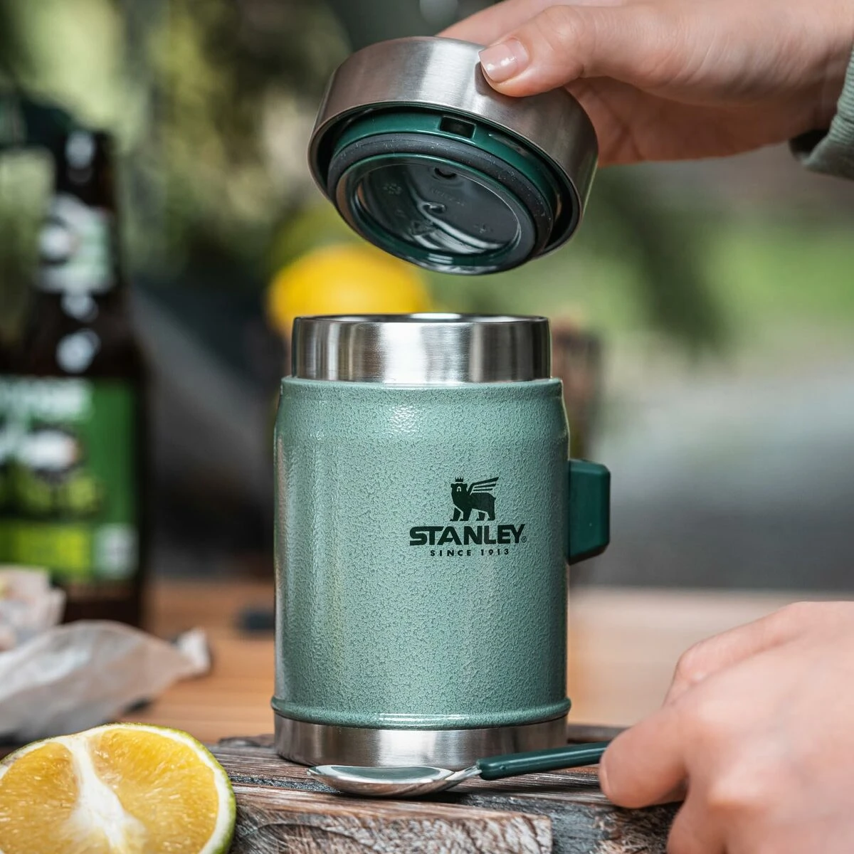 Hand opening a green Stanley insulated mug on blurred background.